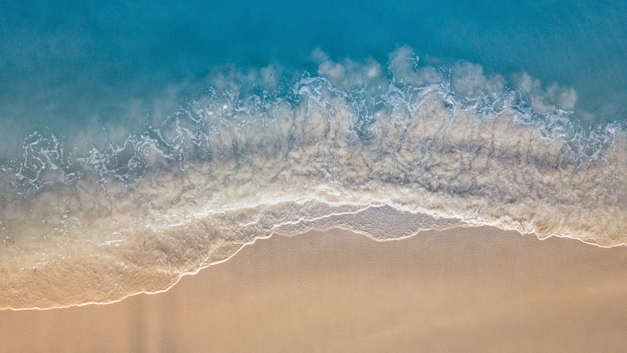 Aerial view looking straight down on a curve of white surf breaking across empty sand