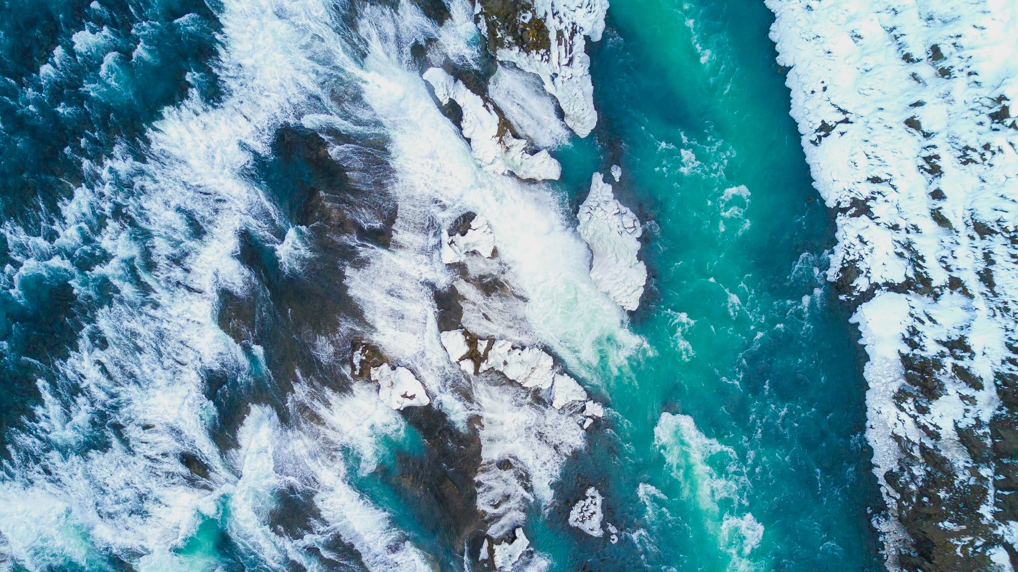 Aerial view looking straight down on a turquoise river running fast between snow-covered rocks
