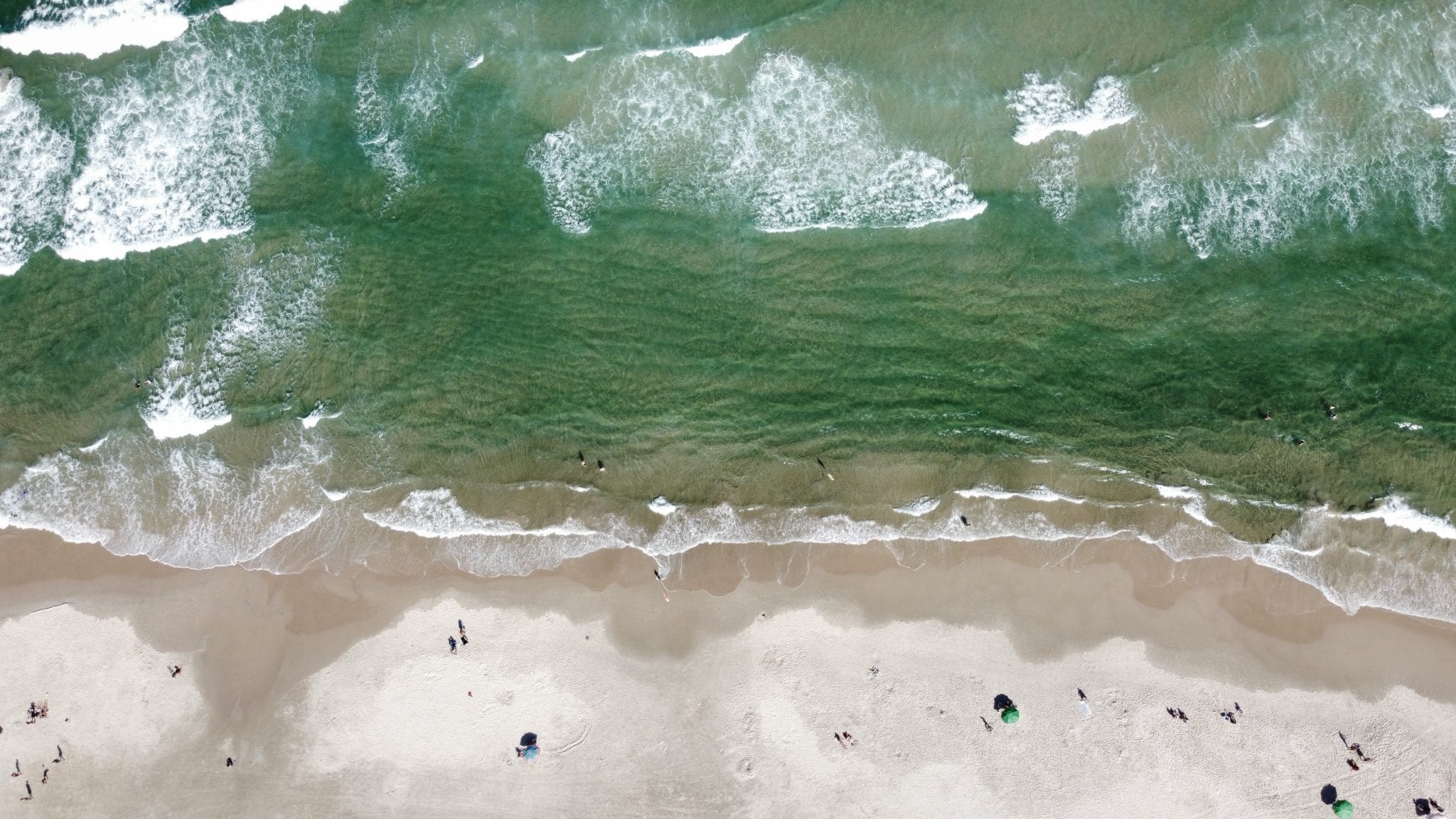Top-down aerial view of beachgoers and umbrellas along a bright green surf line